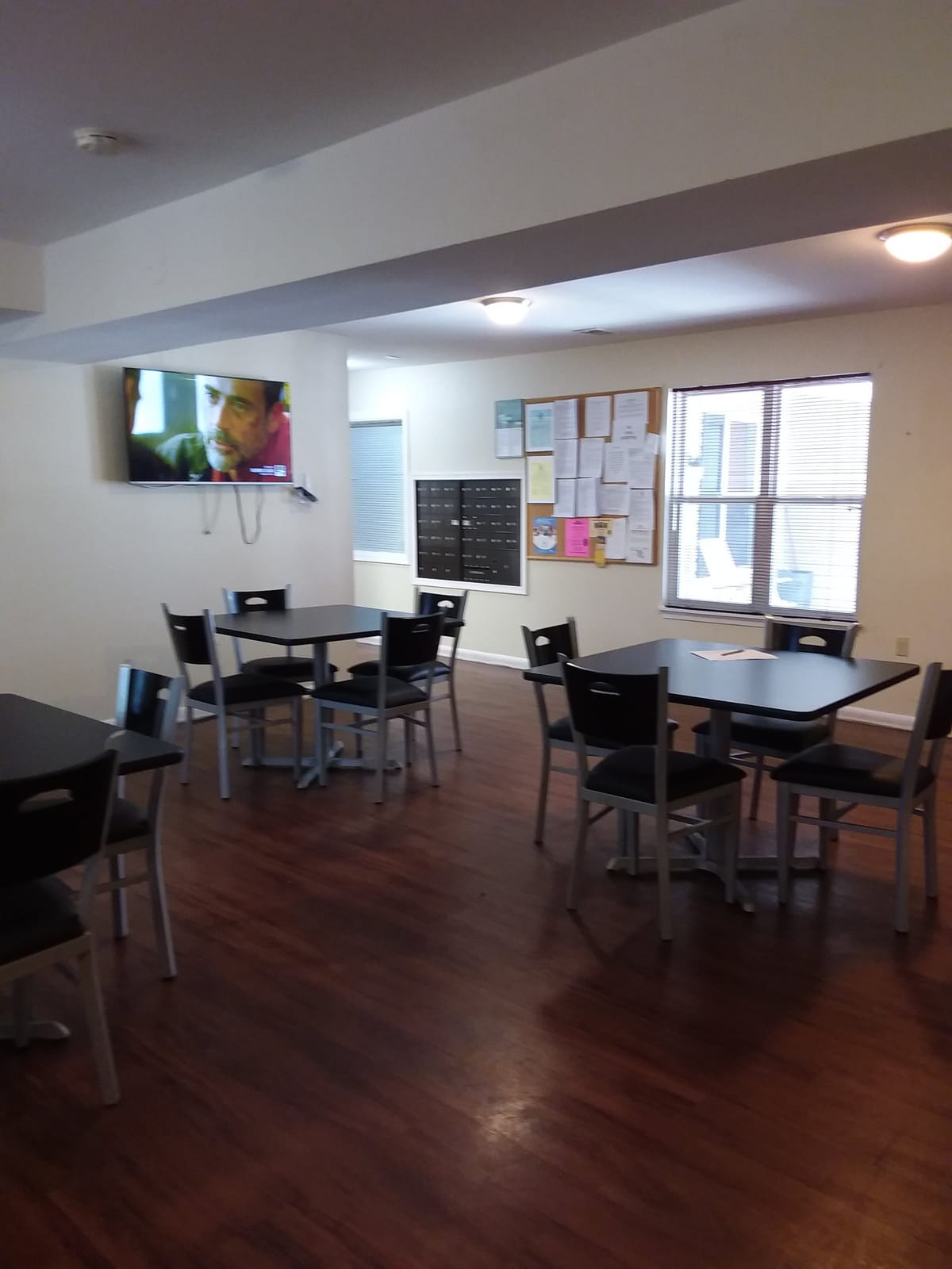A corner of the community room at Indian Head Elderly Apartments with a wall-mounted television, resident mailboxes, a bulletin board, and tables by the windows