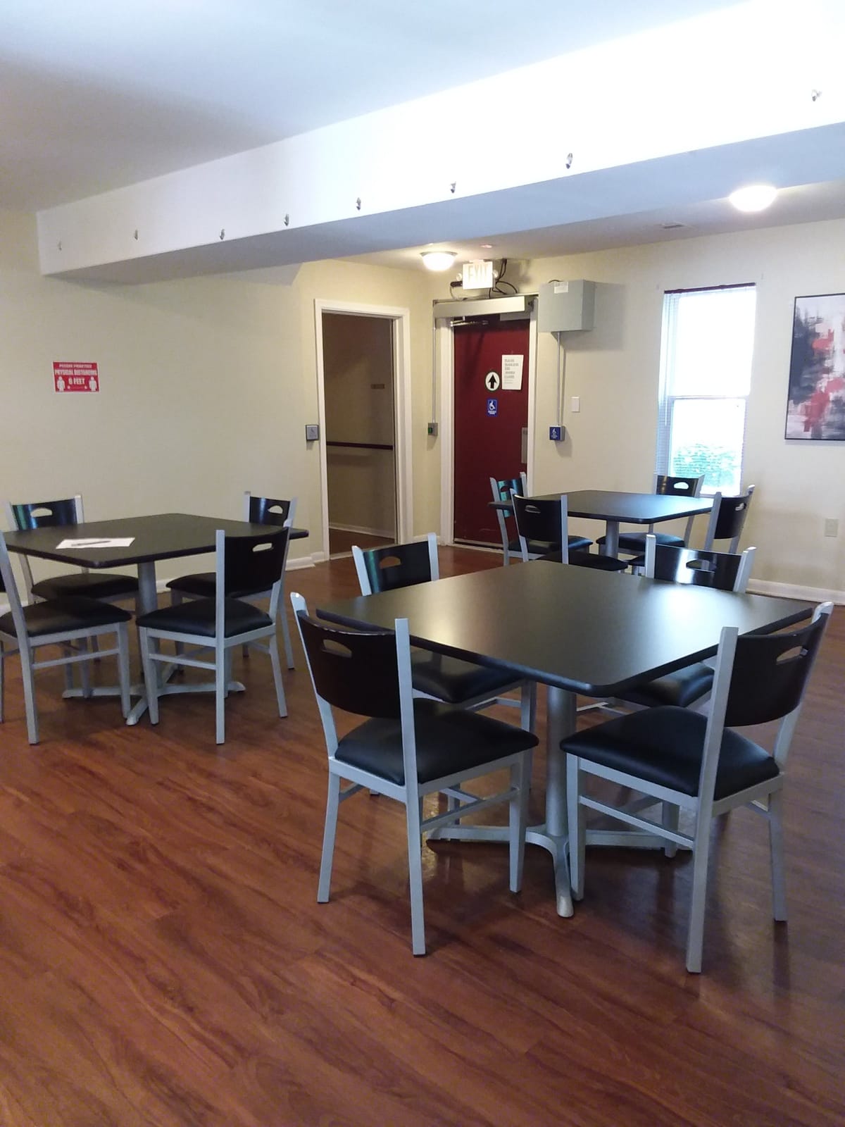 A wider view of the community room at Indian Head Elderly Apartments, with several tables and chairs, a window, and doorways at the far end