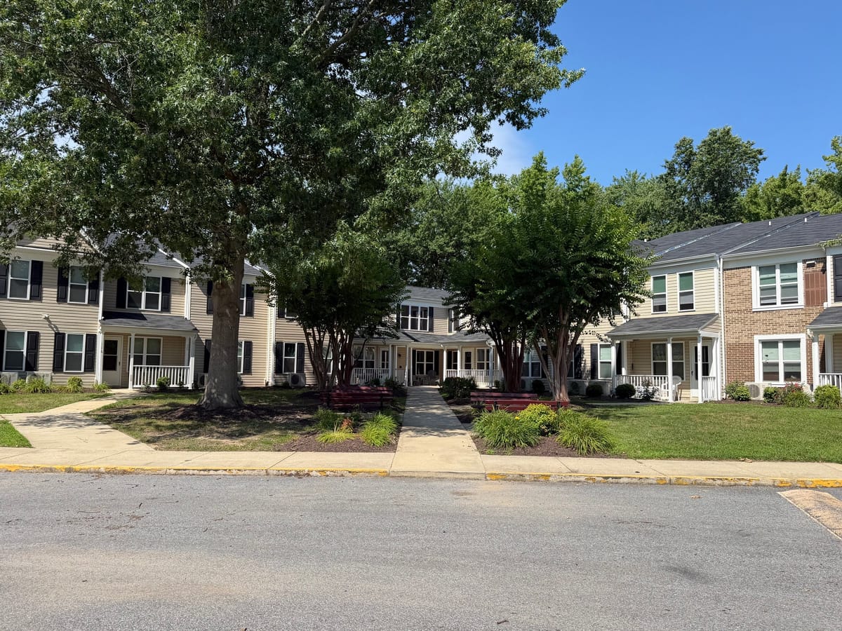 The courtyard at Indian Head Elderly Apartments, with red benches and a concrete walkway running between two rows of two-story tan-sided homes under tall shade trees