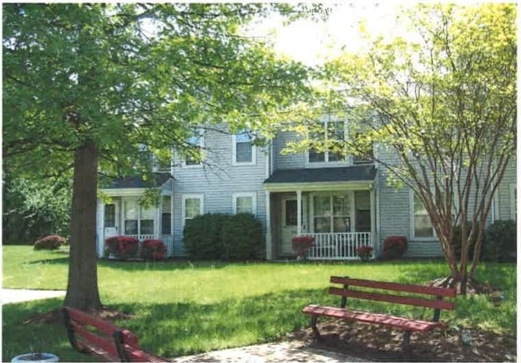 Two-story Indian Head Elderly apartment homes with covered porches, a large shade tree, and a green lawn in spring