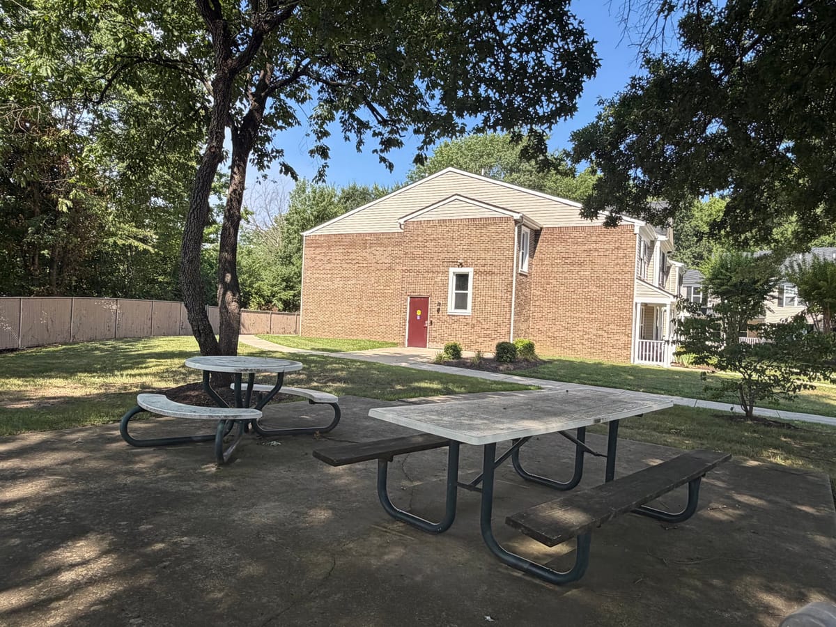 A concrete patio at Indian Head Elderly Apartments with two picnic tables under a shade tree beside the brick community building